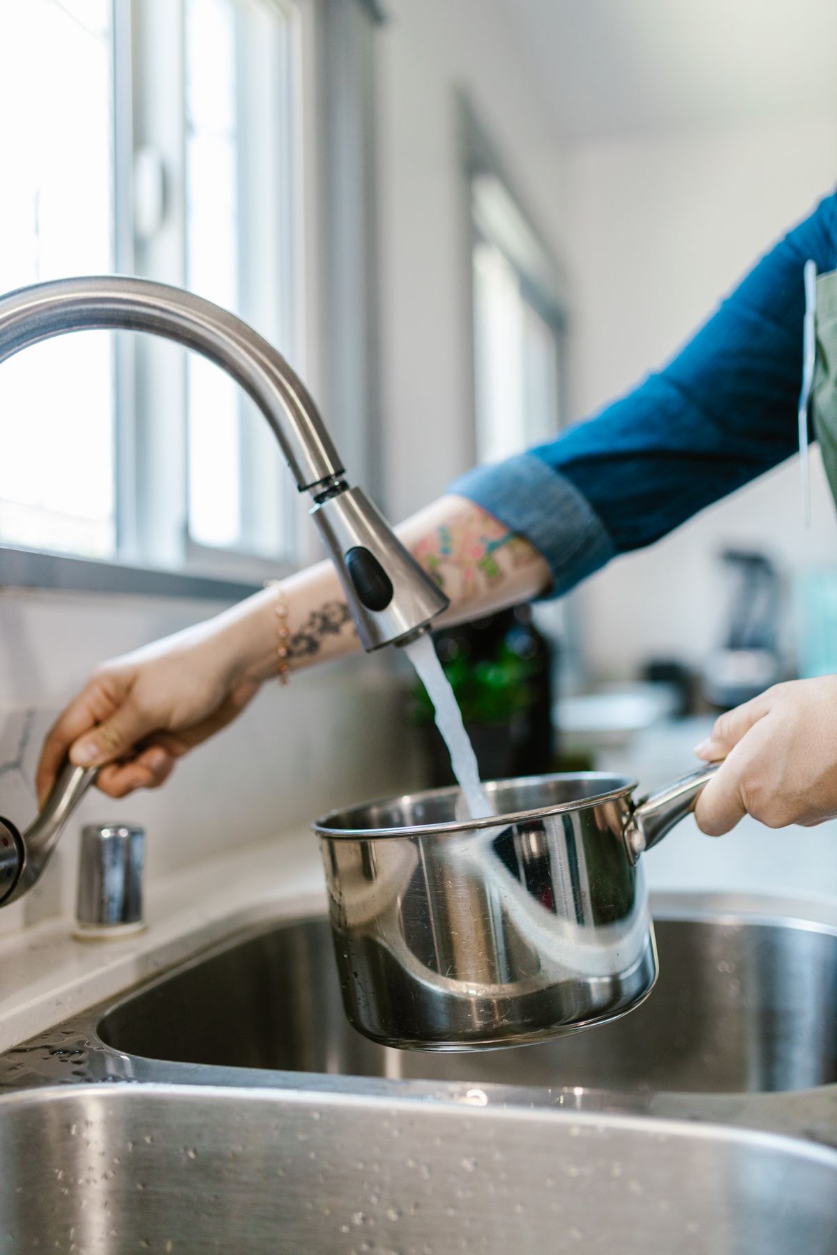 Hand filling a stainless steel saucepan with tap water from a pull-down kitchen faucet over a double sink