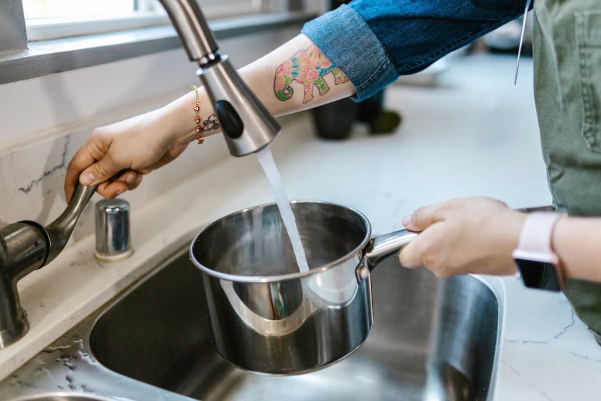 Person filling a stainless steel saucepan at a kitchen faucet, everyday water use that a softener's reserve capacity must cover until regeneration