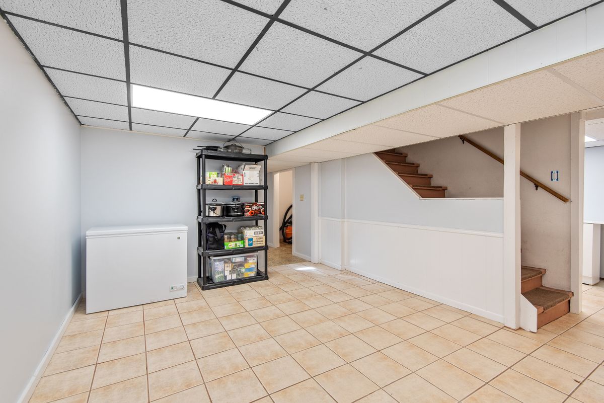 Basement utility space with tiled floor, storage shelving and a staircase, the kind of room near the pressure tank where a well softener, drain and outlet have to line up