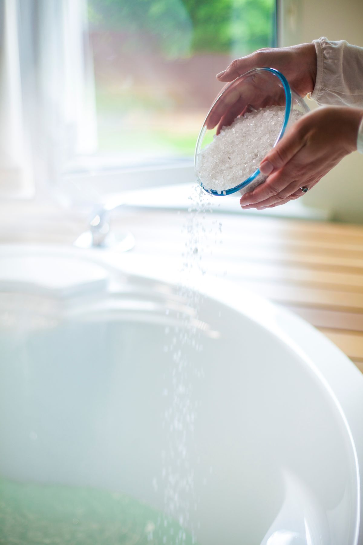 Hands pouring coarse sea salt into a water-filled tub, the same relationship between salt and standing water that governs the level in a brine tank