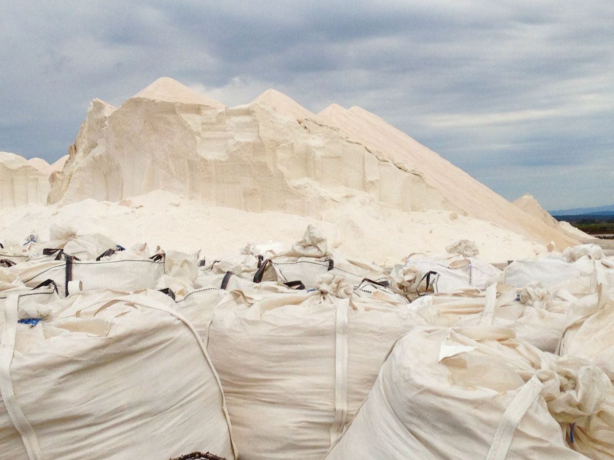 Sacks of salt stacked beside harvested salt piles at an outdoor production site, the bagged supply that a household brine tank is refilled from