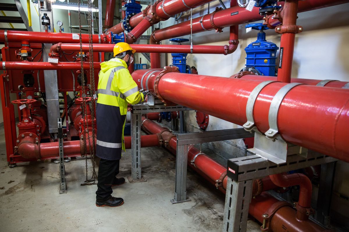 Engineer in safety gear inspecting a run of industrial piping and valves, the isolation step that has to happen before a brine tank is opened