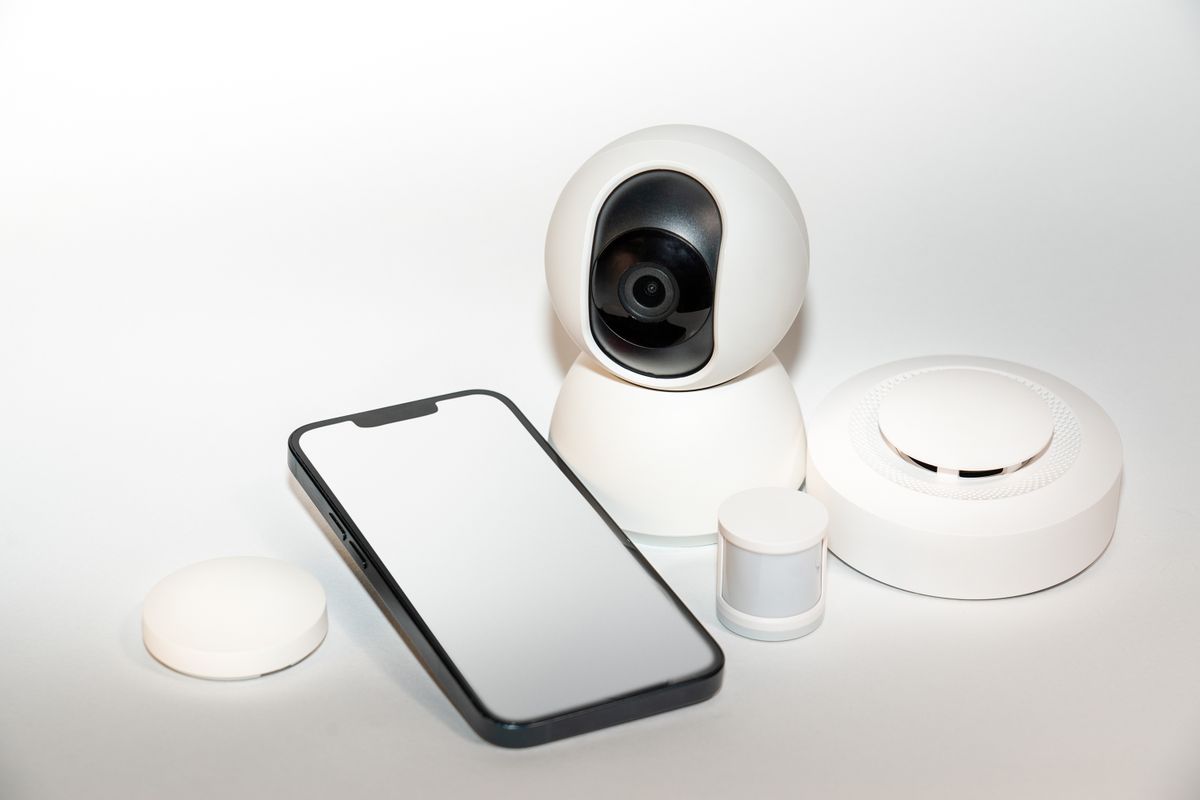 Smartphone lying beside a set of white smart-home sensors and a camera, the connected devices an app-based EcoWater softener alert joins