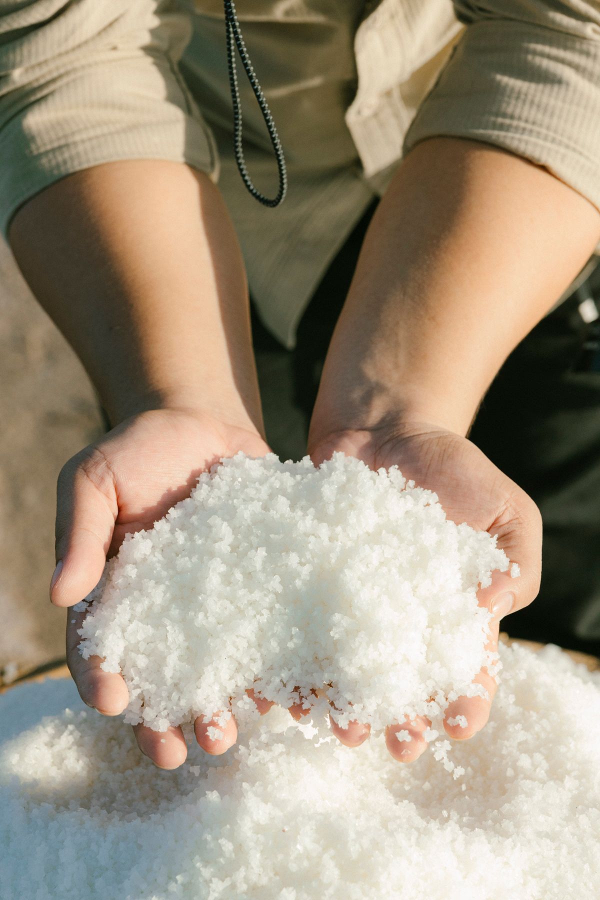 Two cupped hands holding a heap of coarse white salt crystals in sunlight, the salt that should be dissolving out of a softener brine tank but is not