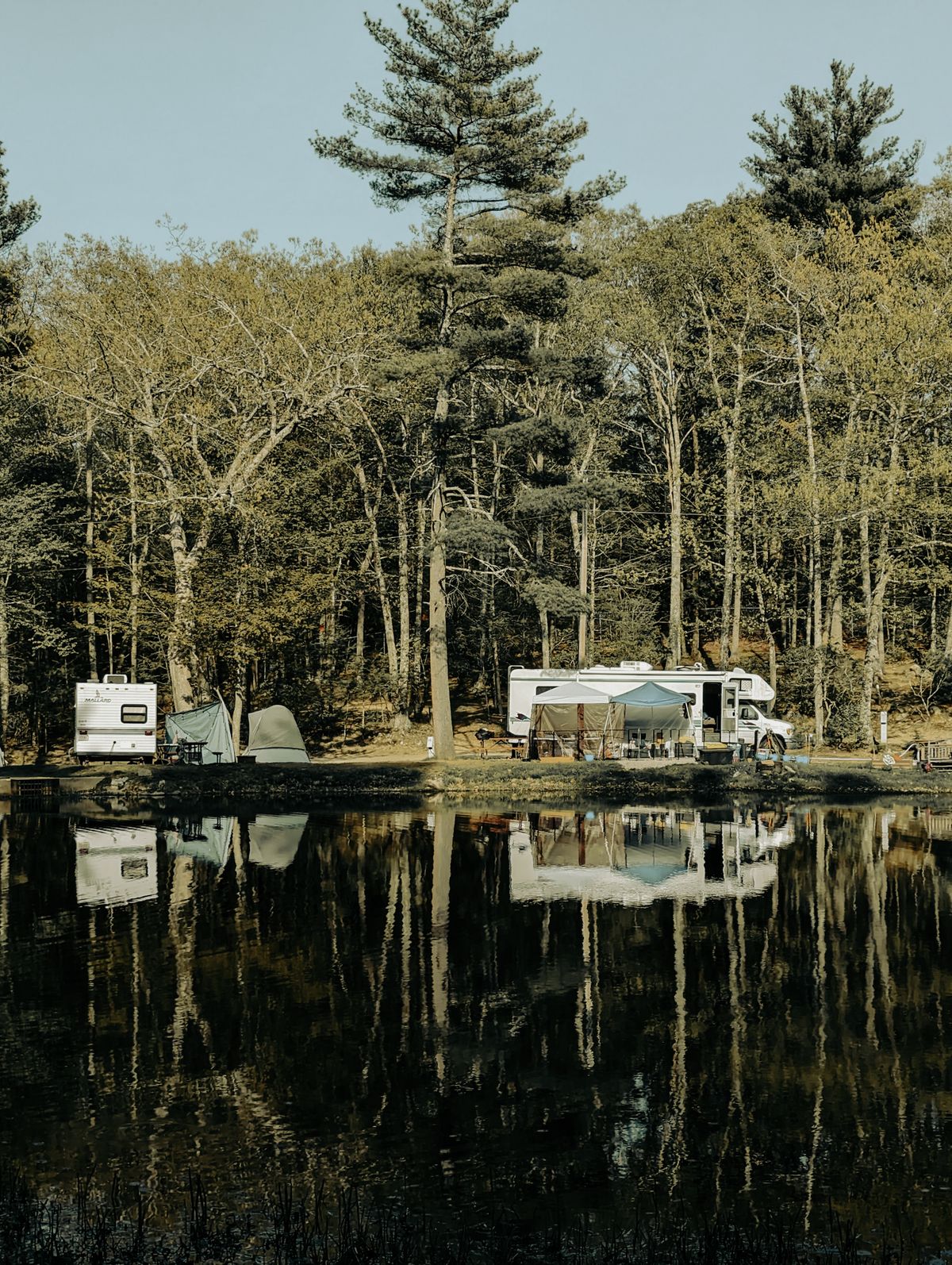 Several RVs and tents camped beside a calm forest lake; each new campground on a route can supply water of a different hardness