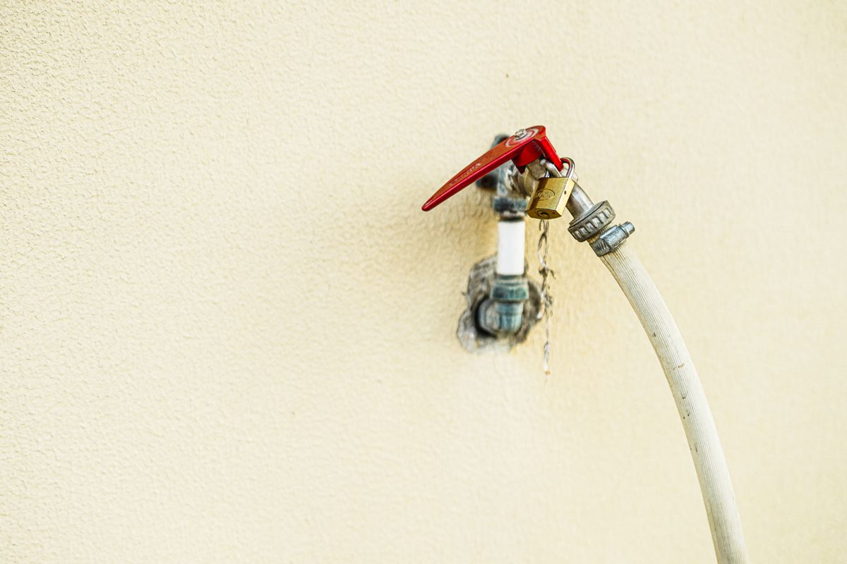 Outdoor hose bib with a red handle and a white garden hose on a cream wall, a tap that is usually left unsoftened on purpose
