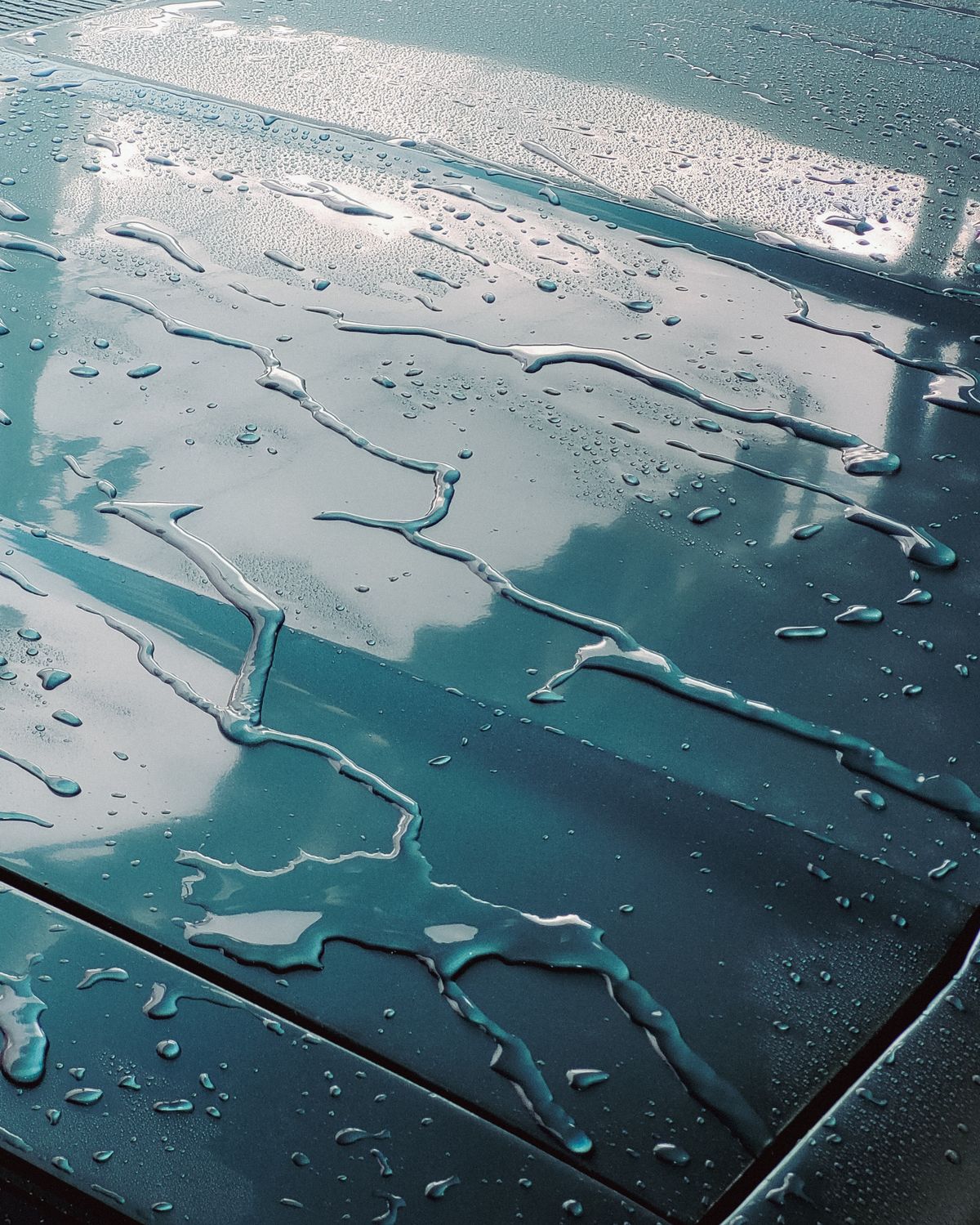 Close-up of water droplets beading on a car hood reflecting clouds and sky, the drops that dry into hard water spots on car paint