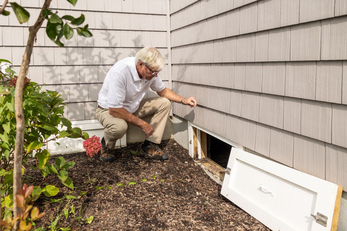 Home inspector crouching beside an open crawl space access door at the base of a house wall, showing how low and tight a crawl space entrance is