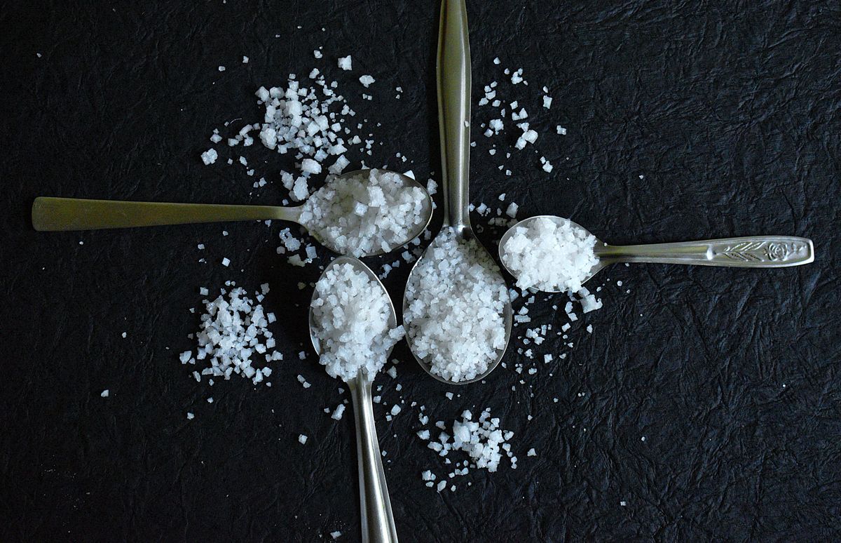 Coarse white salt crystals heaped on four metal spoons of different sizes on a dark surface, four measured amounts of salt