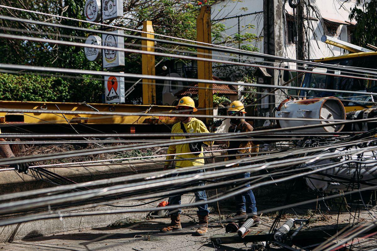 Utility workers in hard hats repairing downed power lines in a street after a storm, cutting power to homes along the street