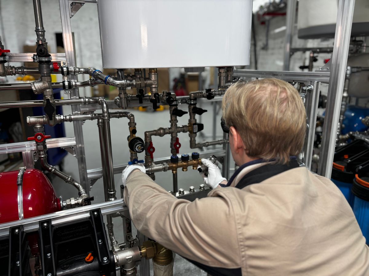 Technician fitting valves and supply piping beneath a white storage water heater tank, with a red expansion vessel and blue filter housings nearby