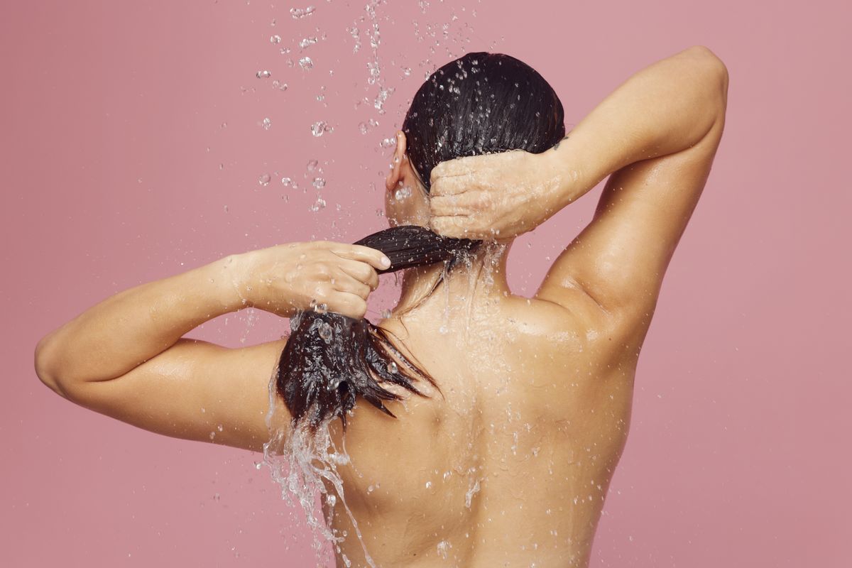 Woman rinsing long dark hair under a running shower against a pink wall, the daily wash that hard water and chlorine both affect