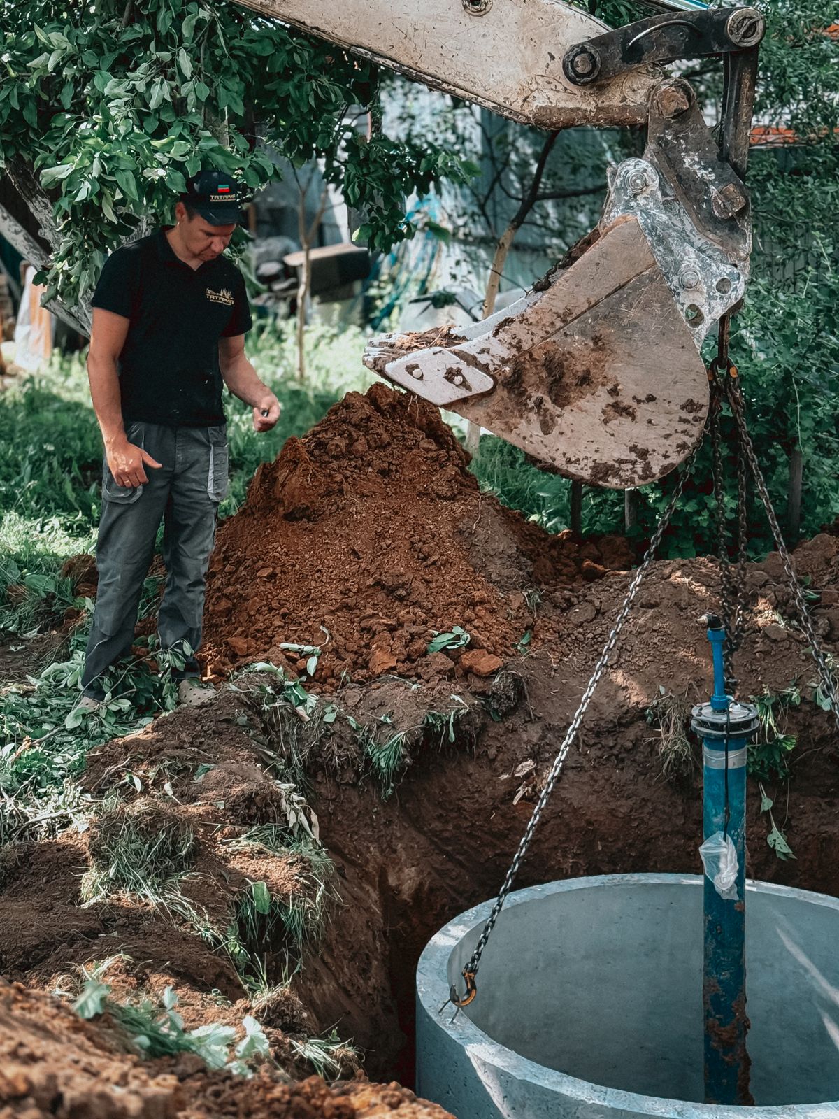 Excavator lowering a concrete ring around a blue well casing during well construction, one of the jobs after which a well is shock chlorinated
