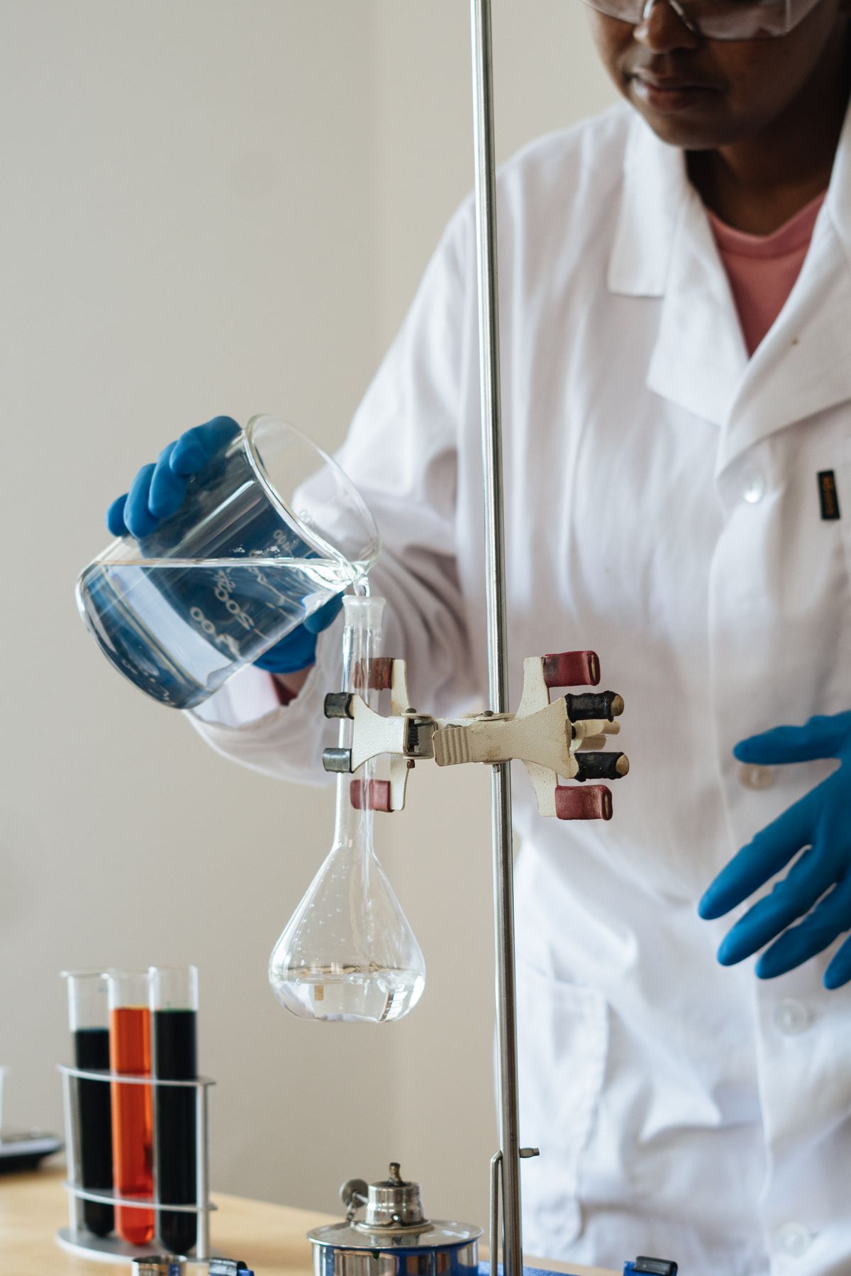 Laboratory technician in goggles and blue gloves pouring a water sample into a clamped flask during product testing
