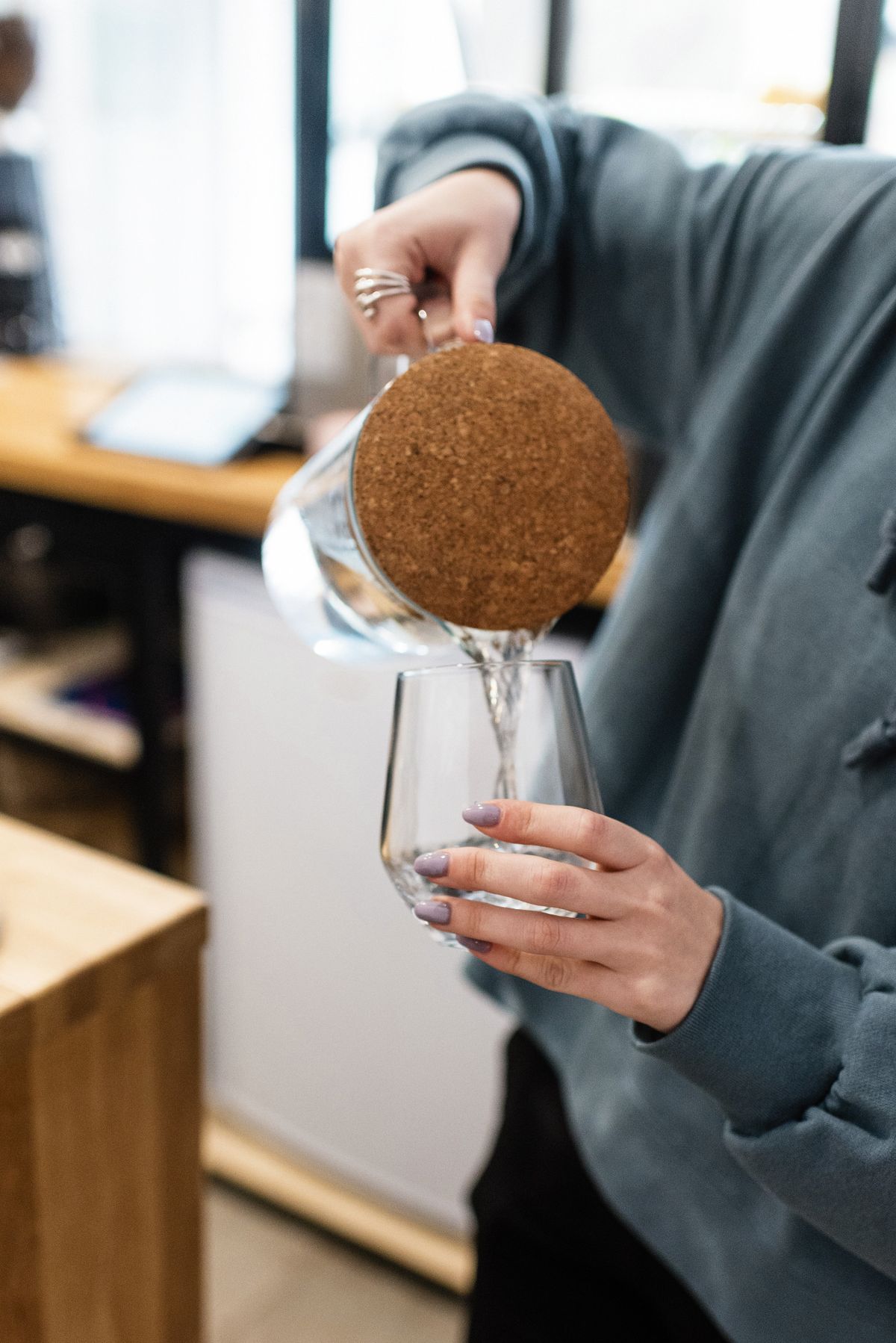 Person pouring clear water from a cork-lidded glass carafe into a drinking glass in a kitchen
