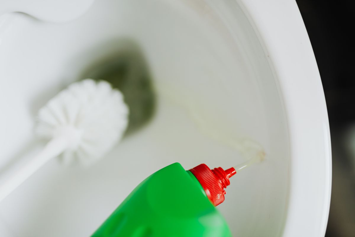 Cleaning liquid being poured from a bottle into a toilet bowl beside a toilet brush