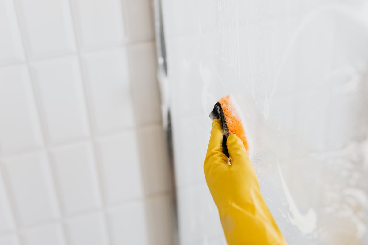 Gloved hands scrubbing a shower screen with a sponge, tackling deposits that persist even when a hardness test reads zero