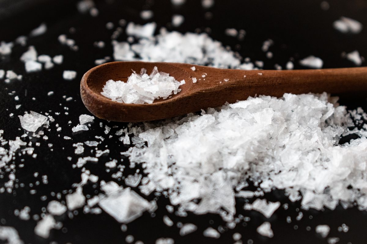 Coarse white salt crystals heaped beside a wooden spoon; dissolved in the brine tank, salt supplies the sodium that regeneration flushes through the resin