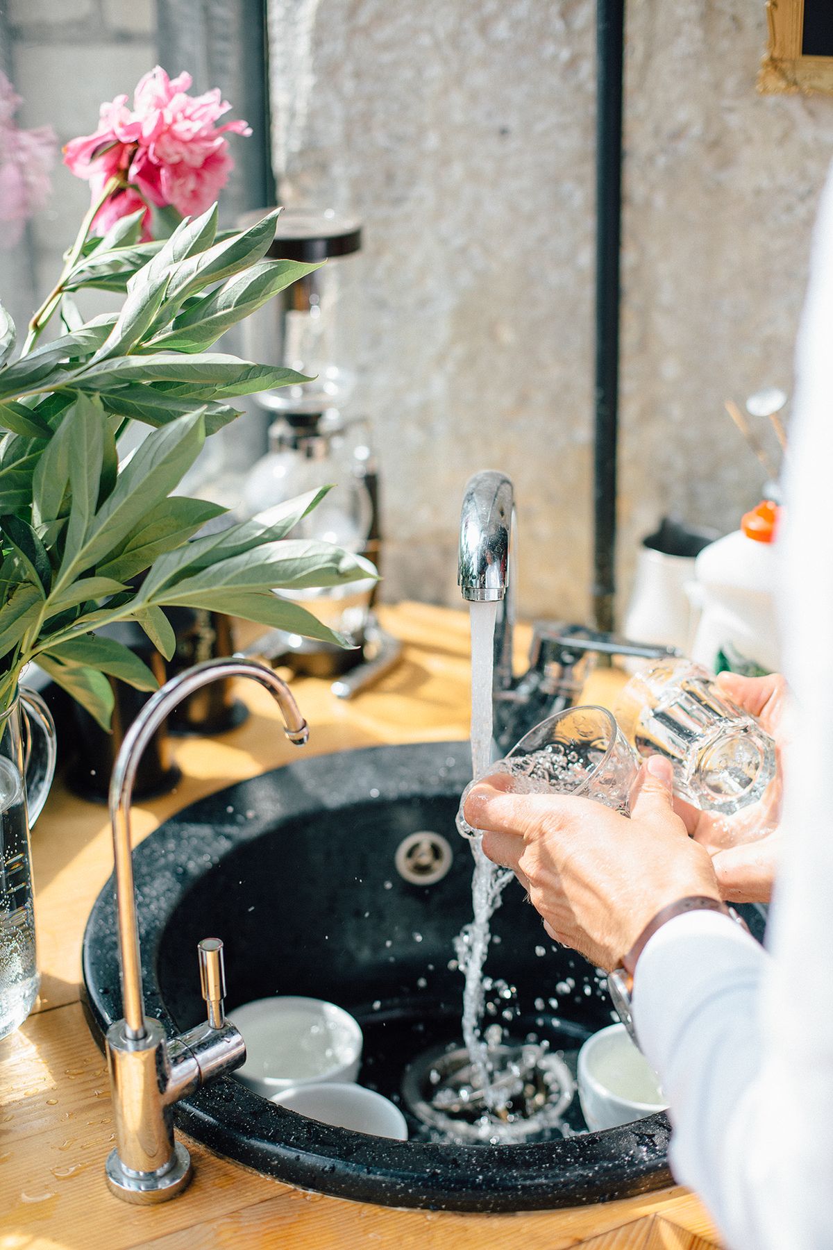 Hands rinsing drinking glasses under a running chrome kitchen faucet into a black sink, where hard water leaves spots on glassware