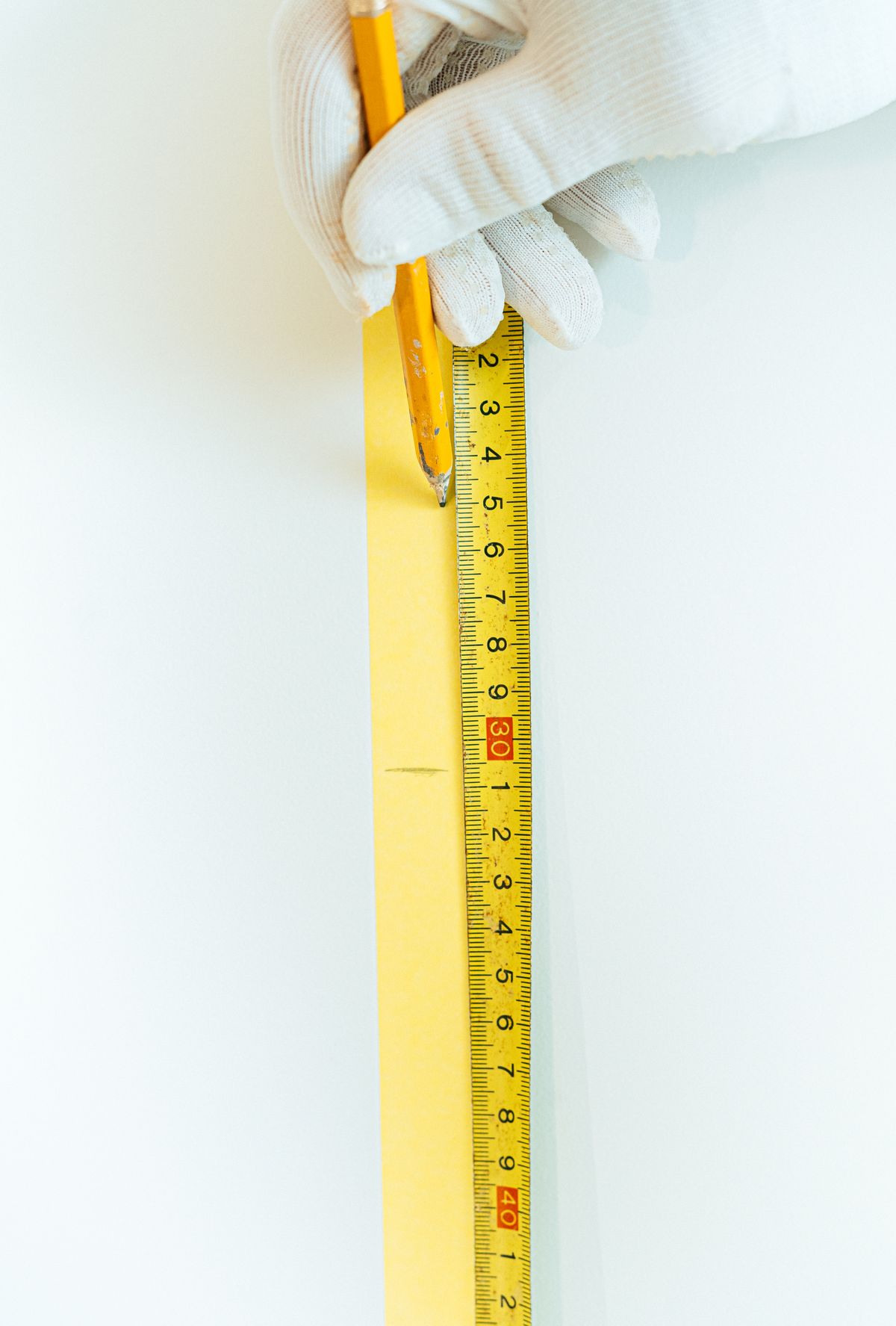Person in a white glove marking a measurement with a pencil against a yellow tape measure held on a wall