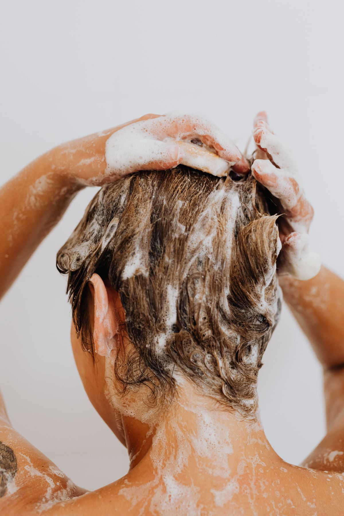 Person washing short brown hair with a thick shampoo lather, seen from behind against a white wall