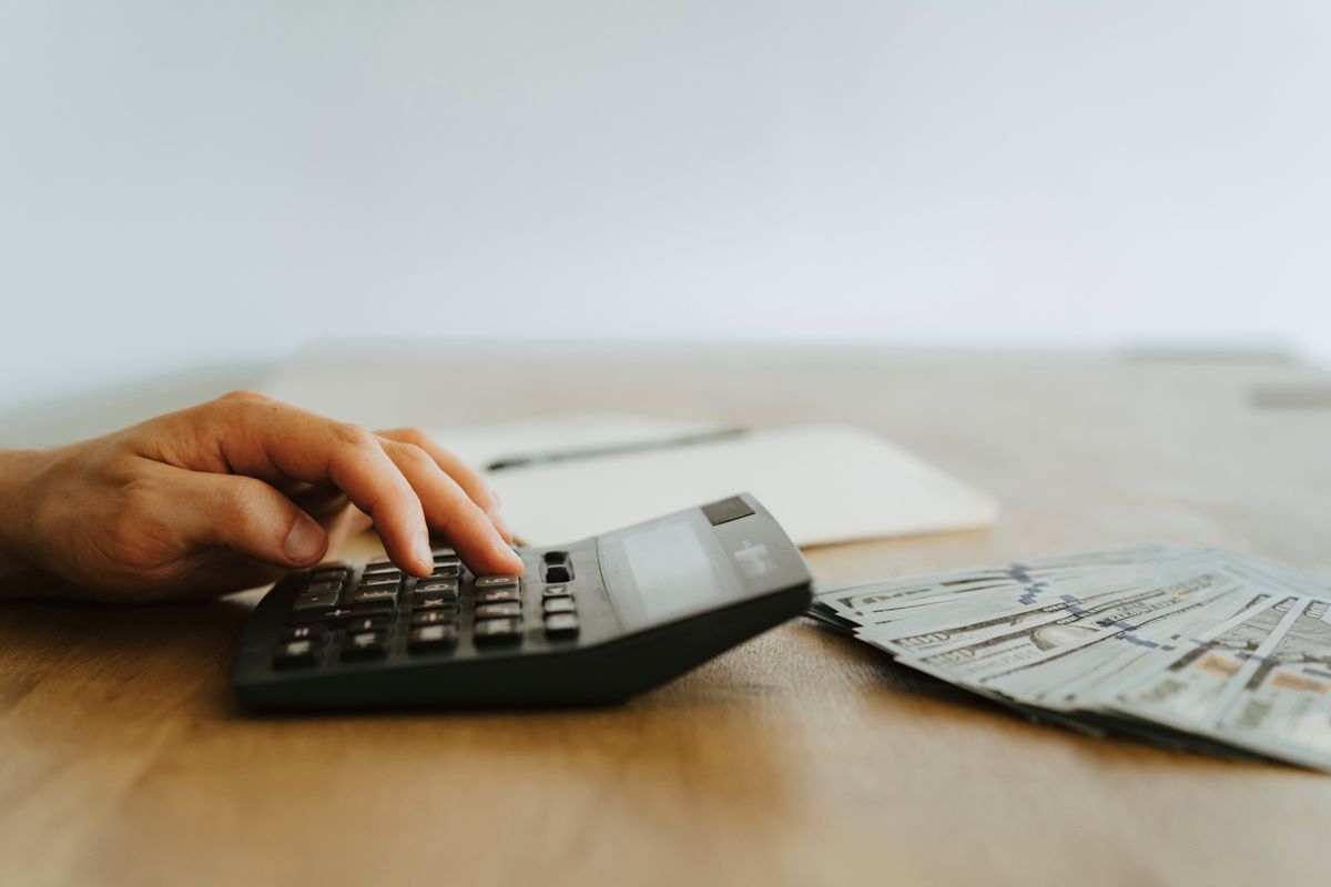 Hand working a calculator on a wooden table beside a fan of dollar bills, adding up water softener prices before choosing a type and size