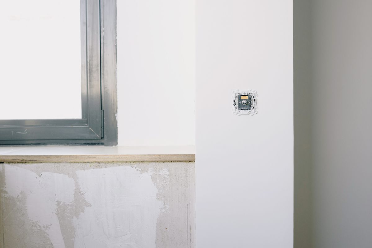 A newly installed electrical outlet box set into a freshly plastered wall beside a window during electrical work in a home