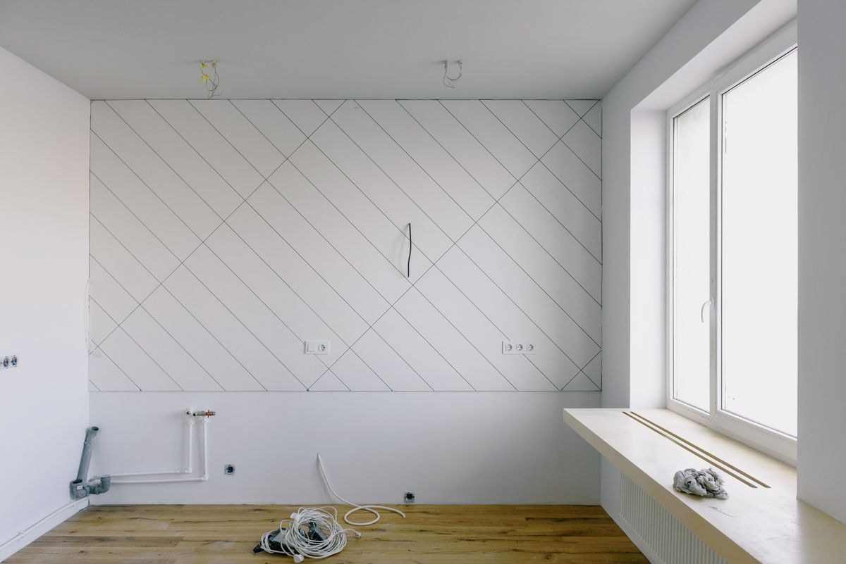 Kitchen mid-remodel with white walls, new supply and drain pipes stubbed out at floor level and electrical cable coiled on the wood floor