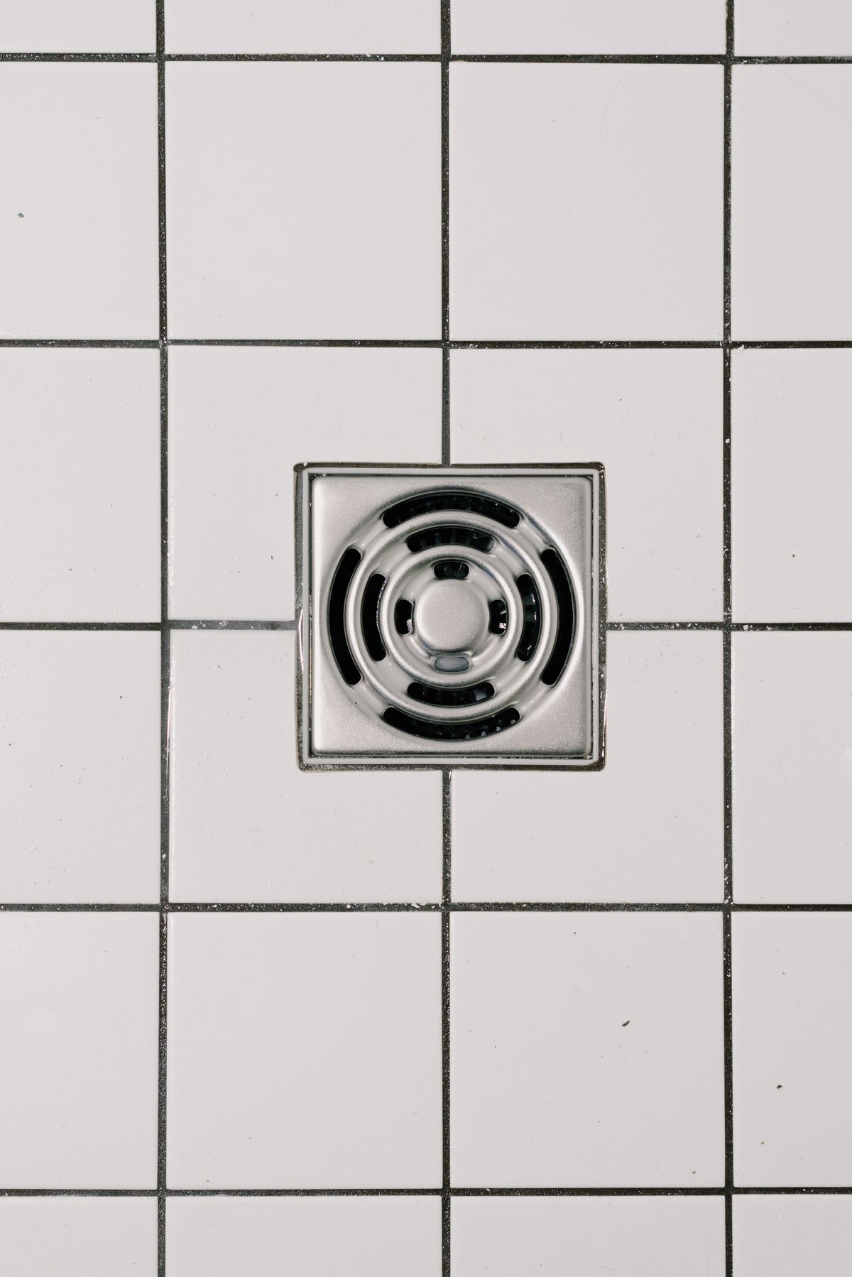 Square stainless steel floor drain set in white tiles, the kind of floor drain where both a softener drain hose and a brine tank overflow line often discharge