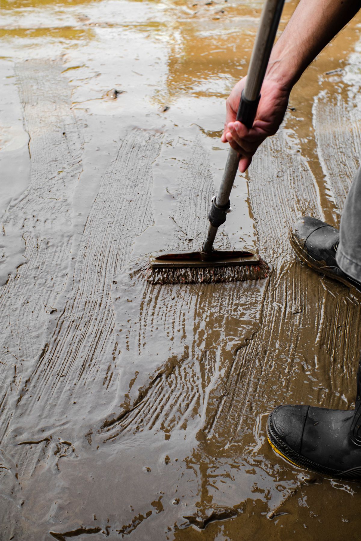 Person scrubbing a wet, muddy floor with a long brush, the scale of cleanup that low-purity salt eventually creates in a brine tank