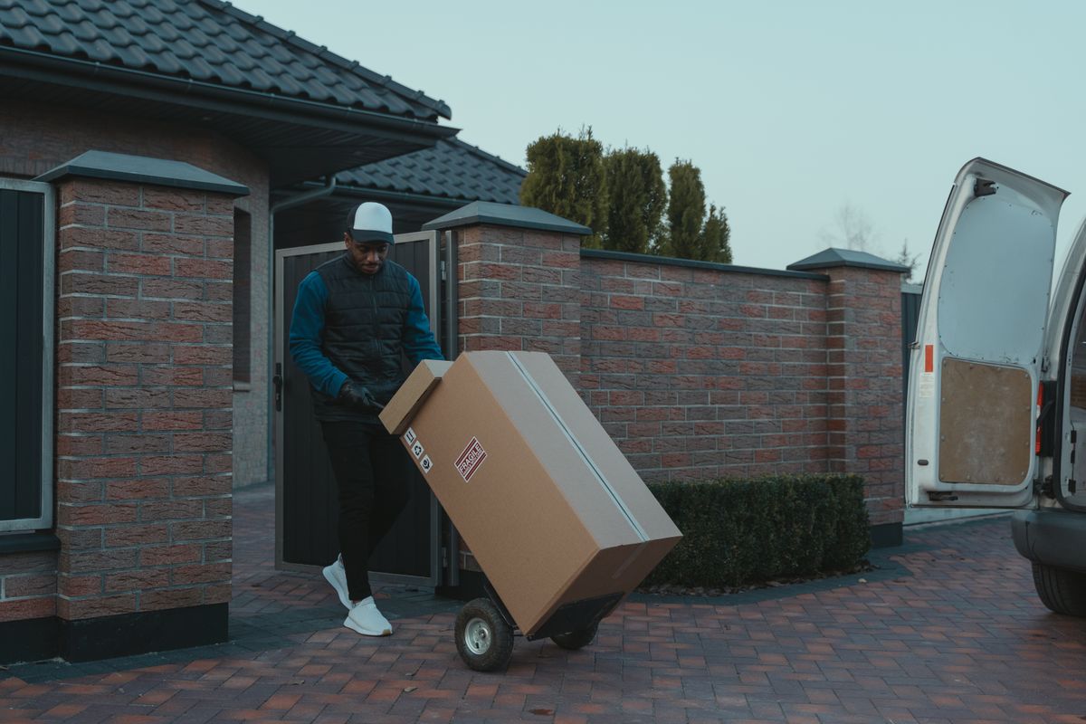 Delivery worker moving a large boxed package on a hand truck along a suburban driveway, the freight a buyer pays when returning a water softener