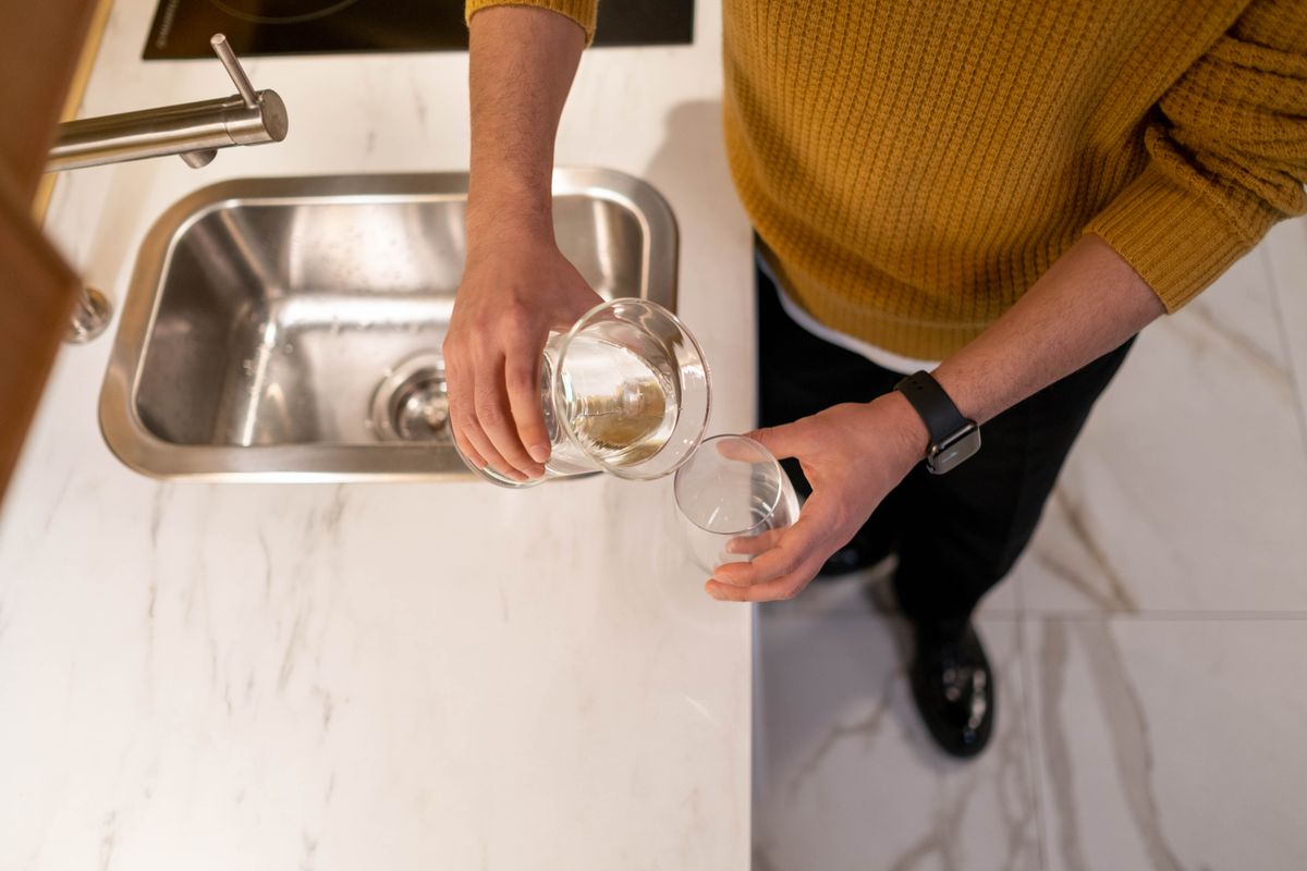 Person pouring water from a glass carafe into a drinking glass beside a stainless steel kitchen sink