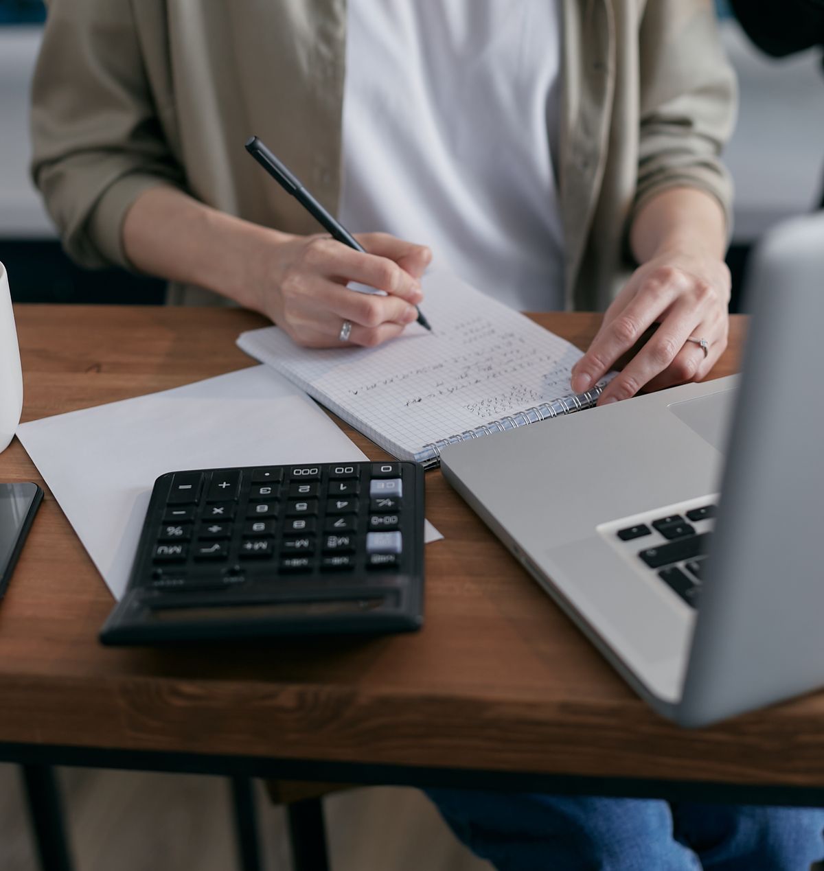 Person writing calculations in a notebook beside a desk calculator and laptop