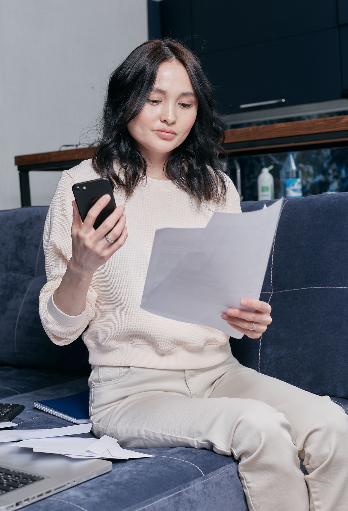 Woman on a sofa checking household bills on her phone and on paper, where a water softener's salt and water costs show up each year