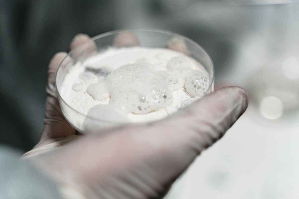 Gloved hand holding a small specimen glass of white powder, representing the dry chemistry of a water softener resin cleaner