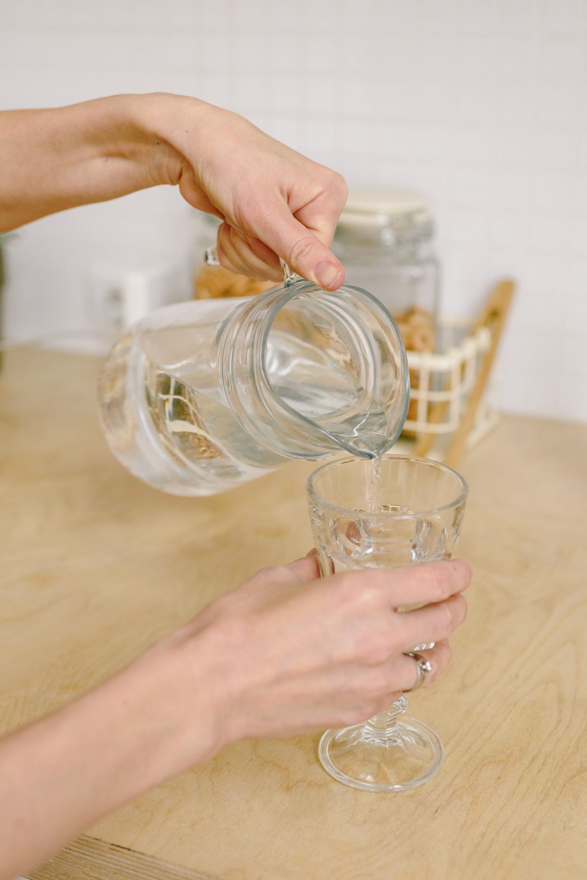 Hands pouring filtered water from a glass jug into a stemmed glass on a wooden kitchen counter