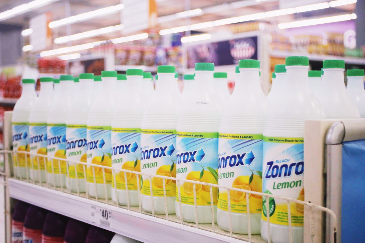 Shelves of household bleach bottles in a supermarket aisle, the same chlorine chemistry that degrades water softener resin at trace levels