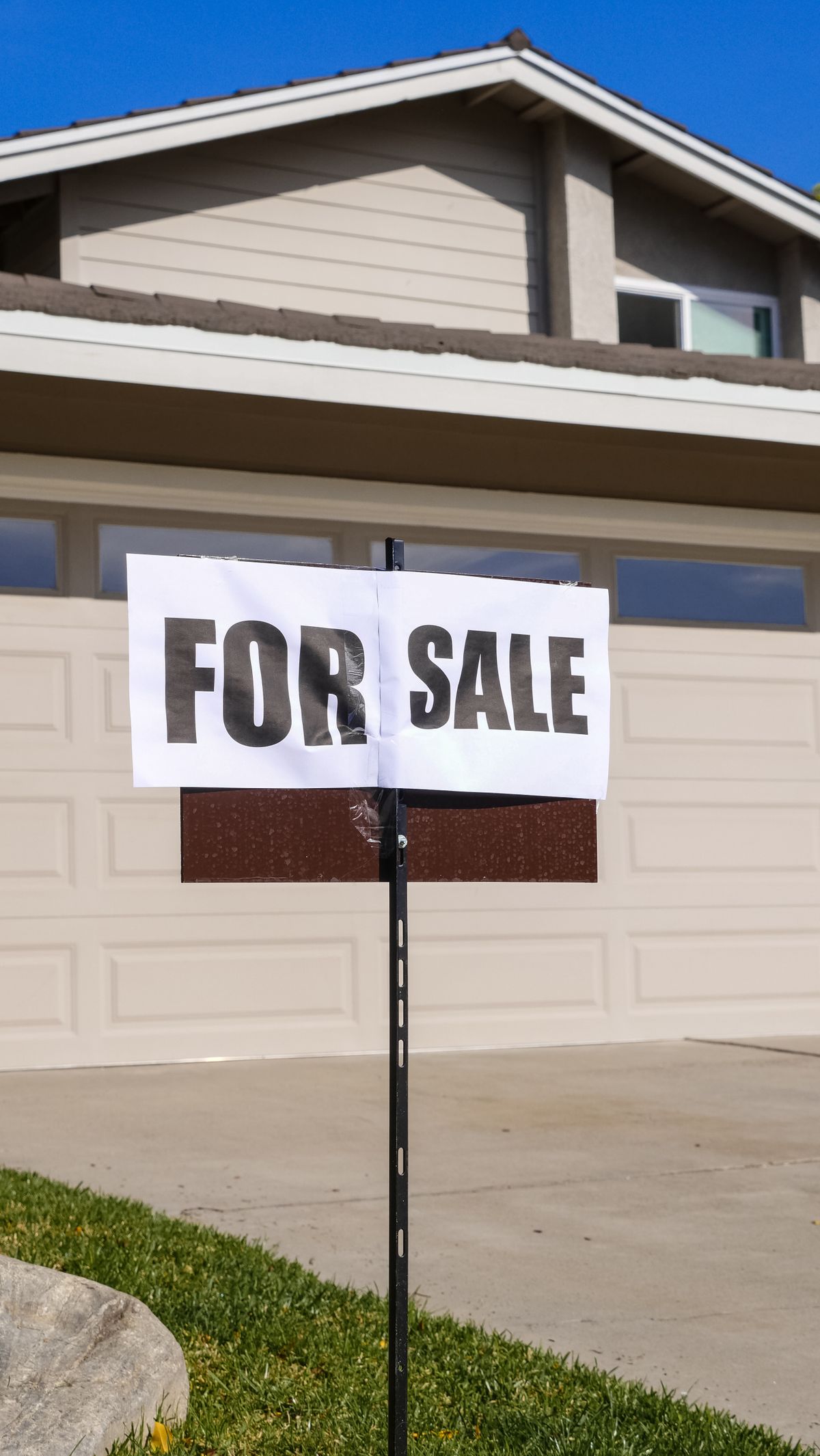 For sale sign on a post in the front yard of a two-story house with an attached garage