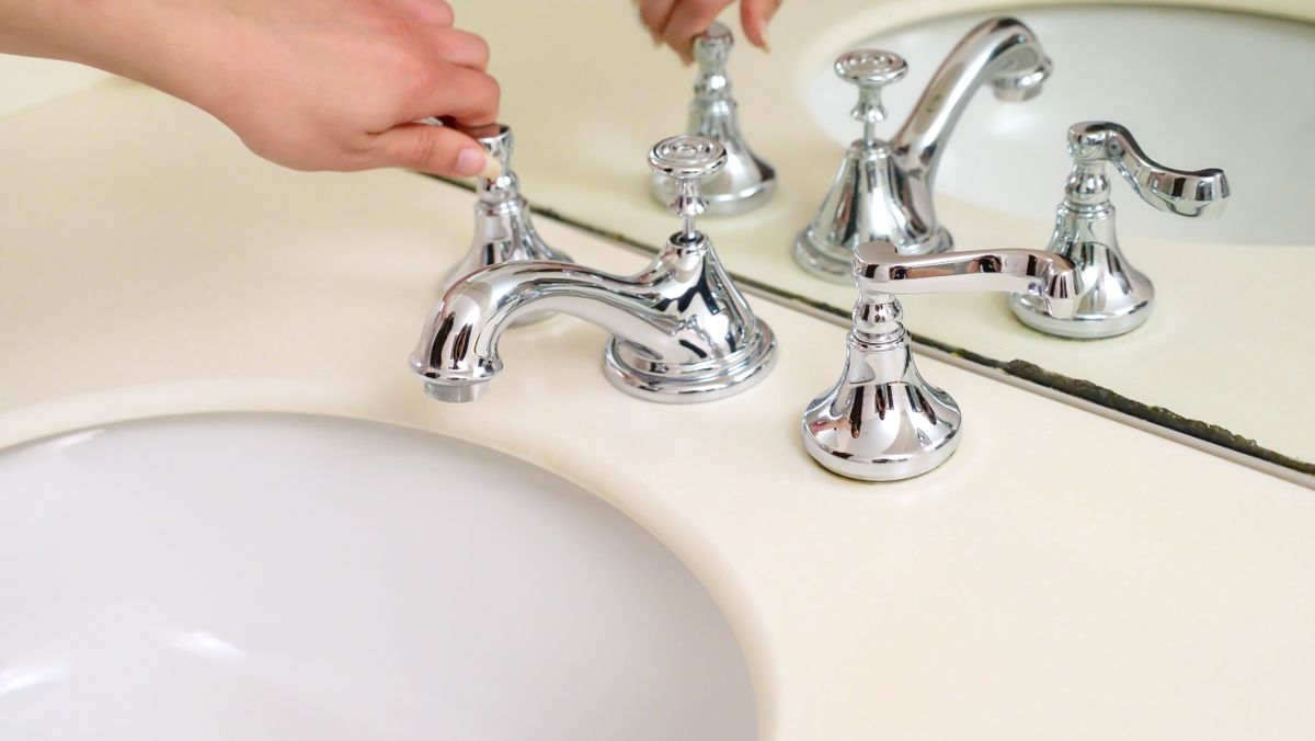 Hands turning the chrome handles of a widespread bathroom faucet, where the cartridge sits inside the valve body