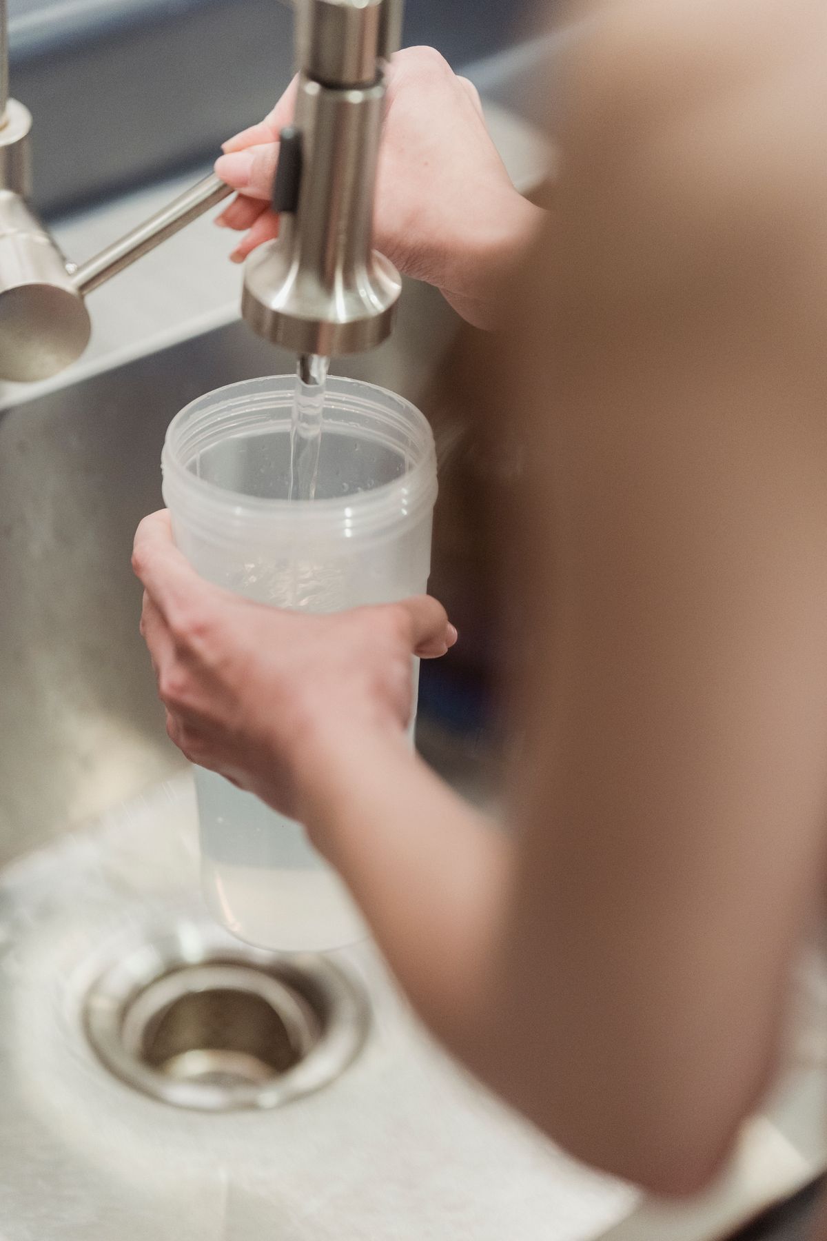 Hand holding a clear cup under a kitchen faucet to collect a flushed cold water sample for a hardness test