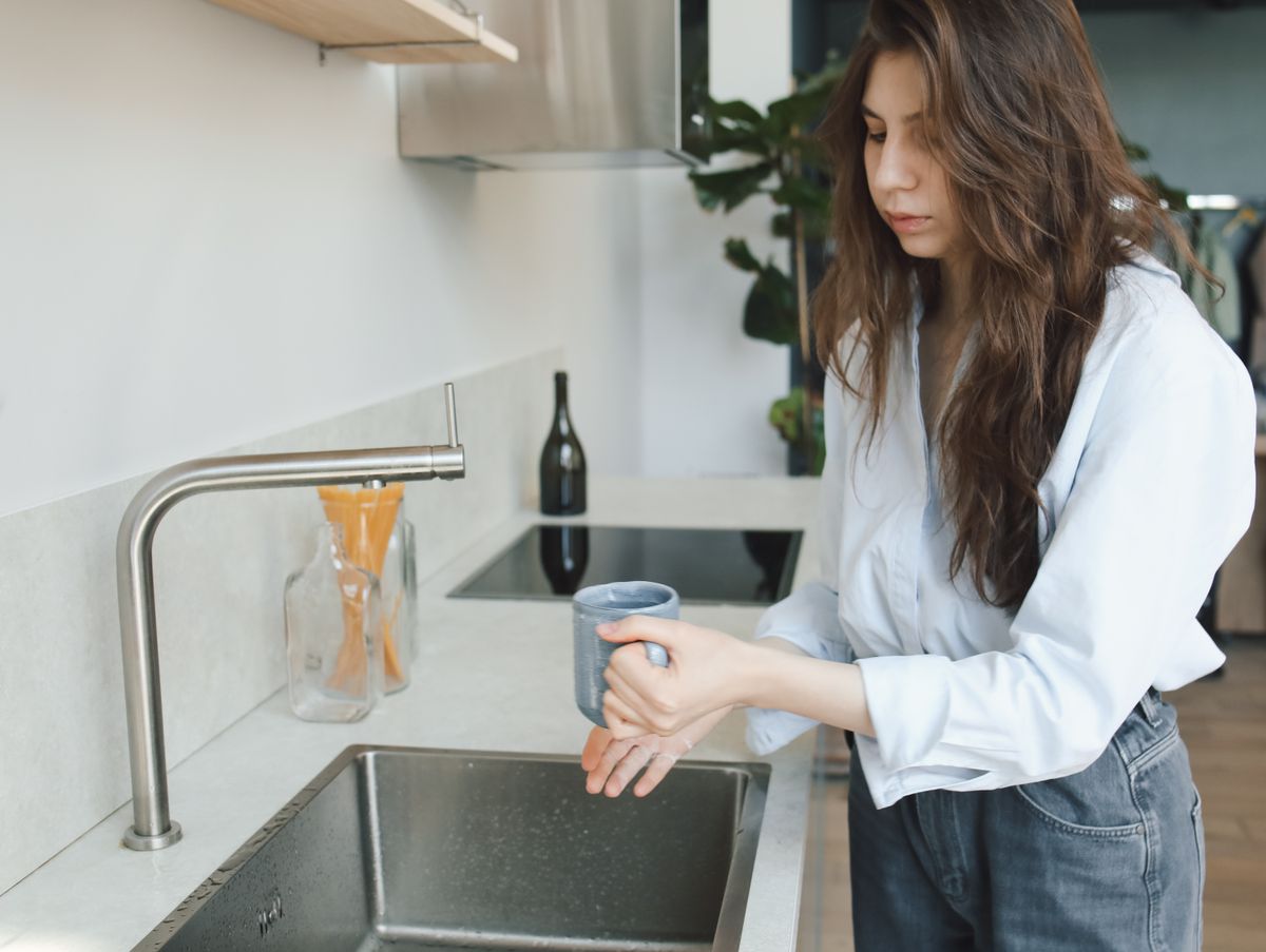 Woman holding a mug at a stainless steel kitchen sink with a tall faucet, the drinking water a whole-house carbon filter treats