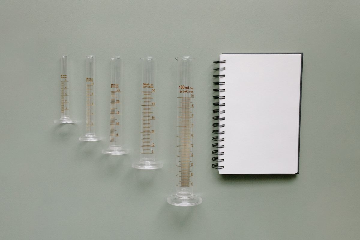 Five glass graduated measuring cylinders lined up beside a blank spiral notebook for logging measurements