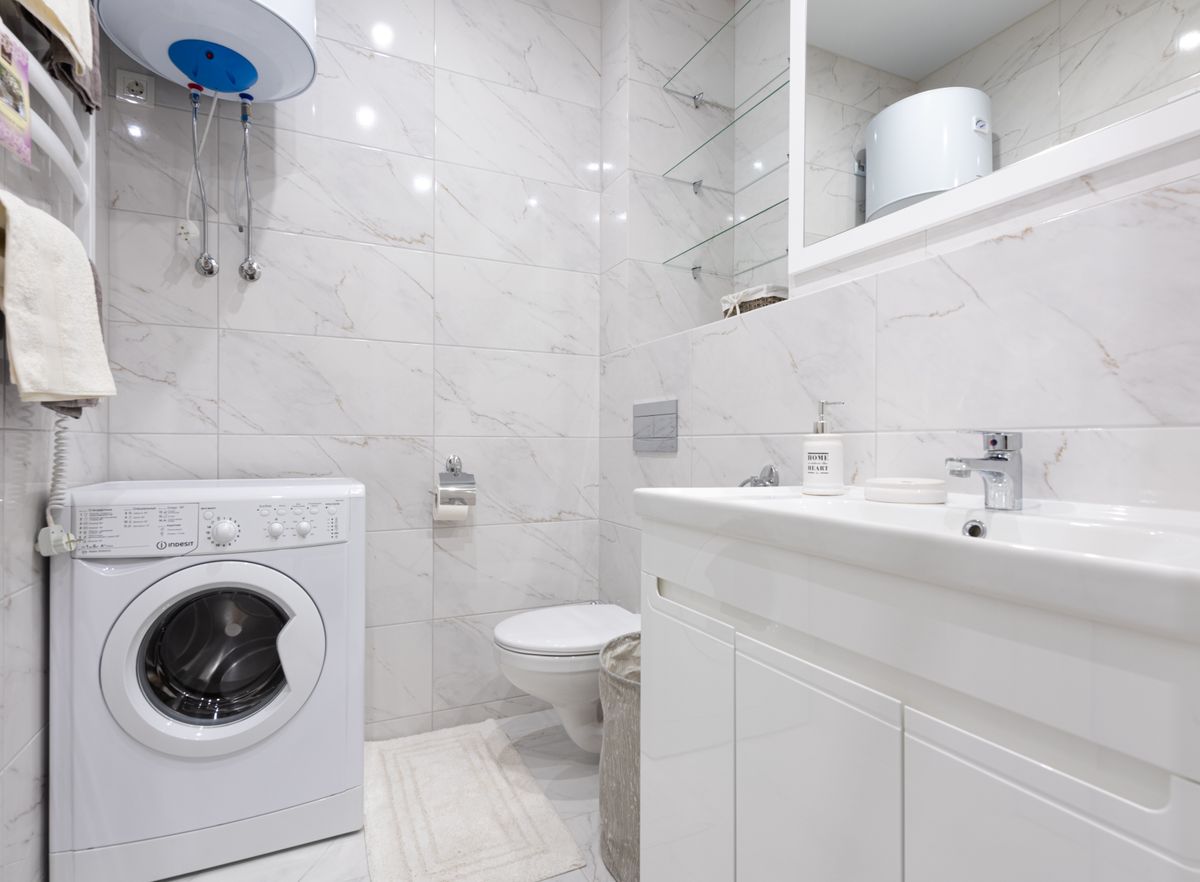 Compact bathroom with a wall-mounted electric water heater above a front-loading washing machine, two appliances a whole house water softener is meant to protect