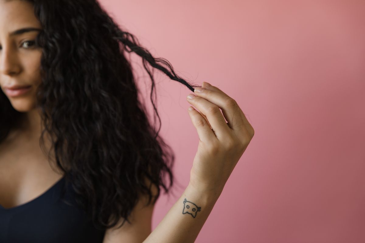 Woman with long curly dark hair holding a strand out between her fingers to check its texture against a pink wall