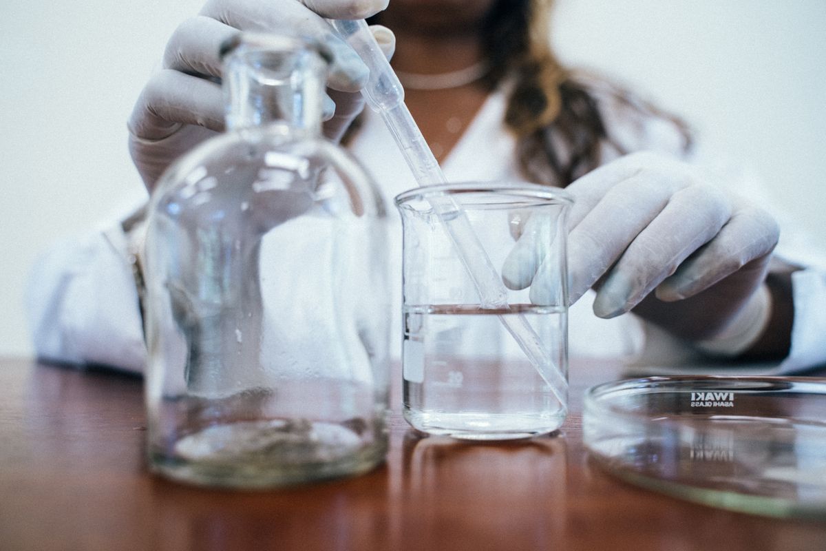 Gloved hands using a pipette to draw a clear water sample from a glass beaker for laboratory testing
