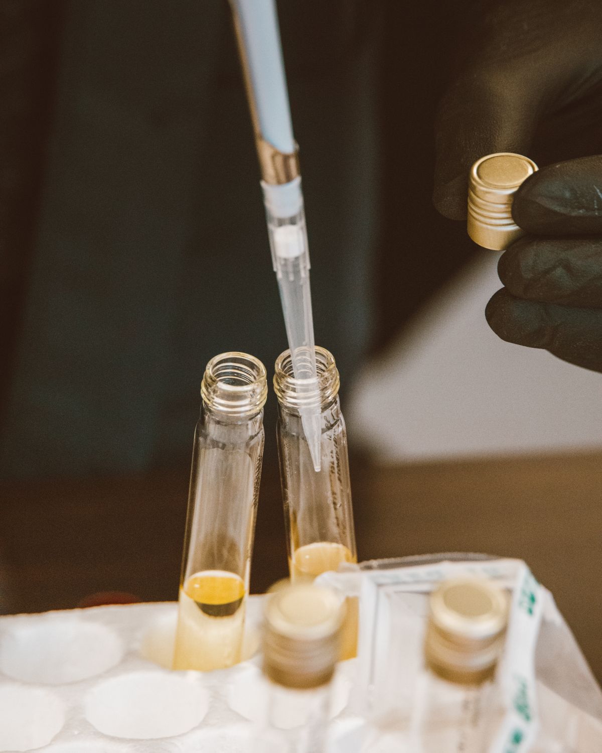 Gloved hand pipetting pale yellow liquid samples into glass vials for laboratory analysis